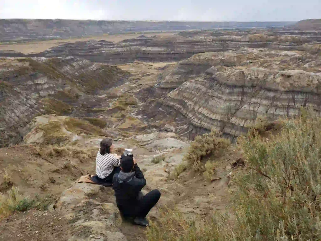 Two people sitting on a rocky cliff overlooking a vast, rugged badlands landscape with layered sedimentary rock formations and sparse vegetation in the foreground