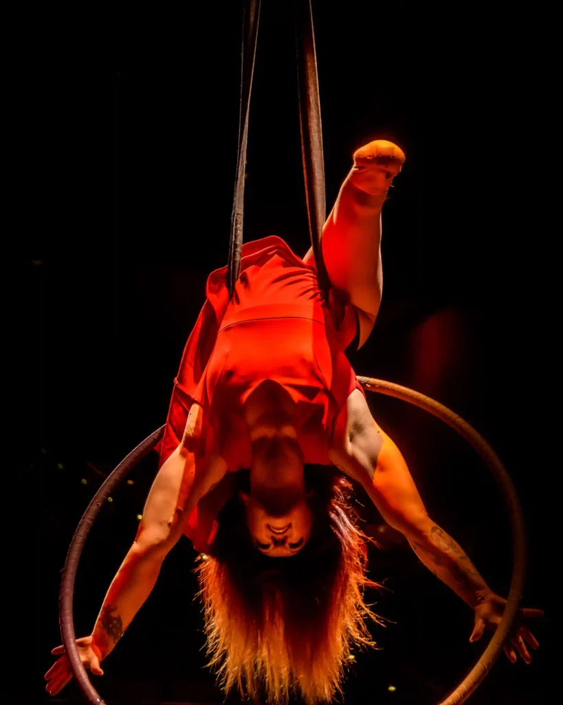 Erin, a white agender human and double below knee amputee, hangs upside down with flaming red hair and a flowing red dress. Erin arches backwards powerfully over the top of an aerial hoop the size of an adult hula hoop that is suspended in the air. Virginia Maria Photography