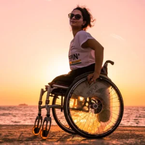 Photo of a confident woman in a wheelchair enjoying a beautiful sunset by the ocean. She is wearing sunglasses, a casual t-shirt, and her wheelchair wheels reflect the warm golden hues of the setting sun.