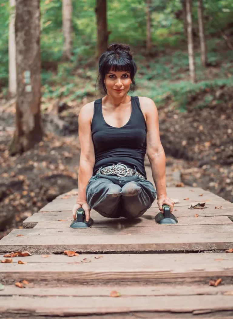 A woman sitting on a wooden bridge in a forest, holding onto push-up bars with her arms straight. She has dark hair tied in a bun with bangs, wearing a black tank top, a decorative belt, and loose jeans. The background shows a lush forest with trees and a natural, serene atmosphere.