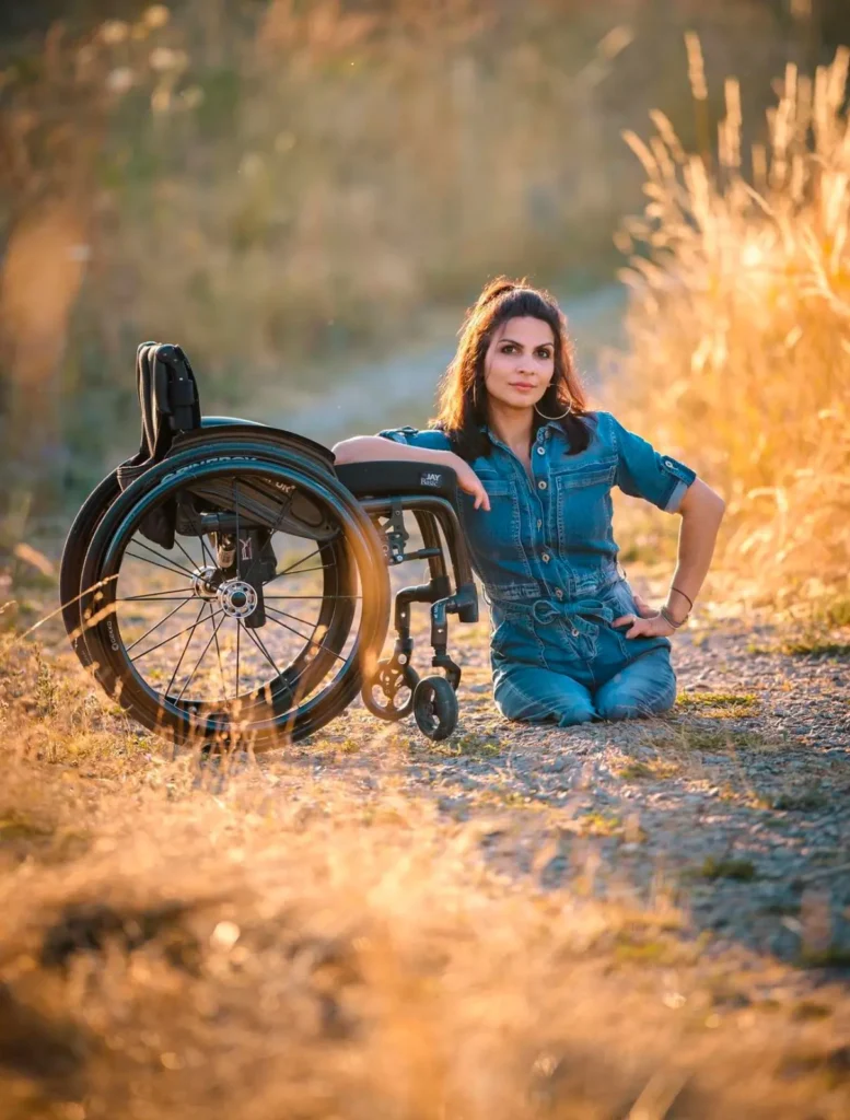Portrait of a woman with a confident expression sitting outdoors in a golden field at sunset, next to a wheelchair. She is wearing a denim jumpsuit, with the sun casting a warm glow on her and the natural surroundings