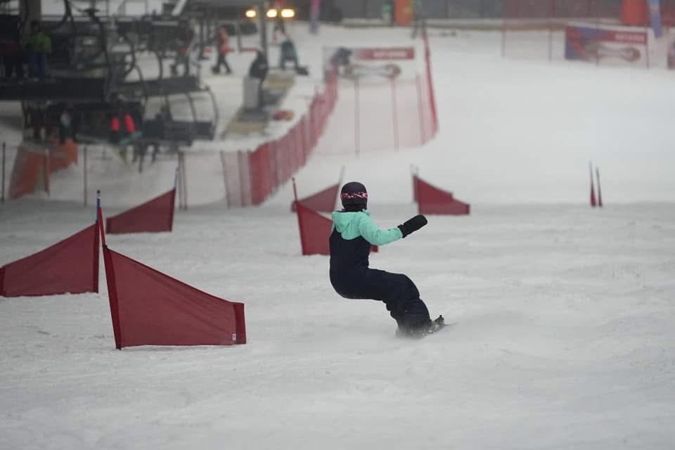 A snowboarder in a light blue and black outfit navigating through a slalom course marked with red flags on a snow-covered slope. The background shows a ski lift and other participants at a winter sports venue.