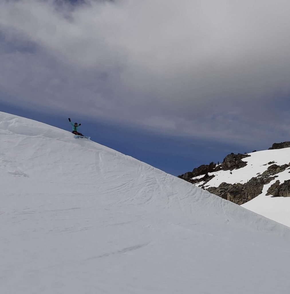 A snowboarder in a green jacket standing at the edge of a snowy slope, raising their arms triumphantly under a cloudy blue sky. Snow-covered mountains and rocky terrain are visible in the background.