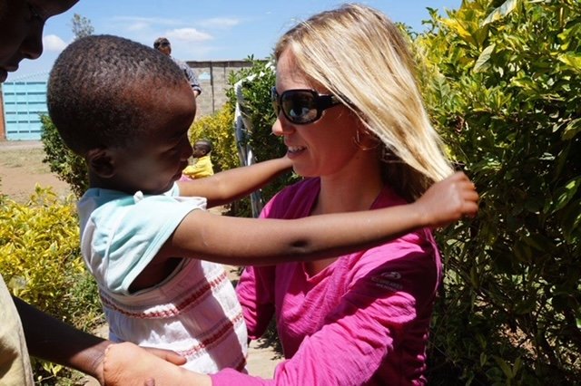 An adult woman wearing sunglasses and a pink shirt interacting warmly with a young child outdoors. The child is reaching out and touching the woman's blonde hair. They are surrounded by greenery and other people can be seen faintly in the background