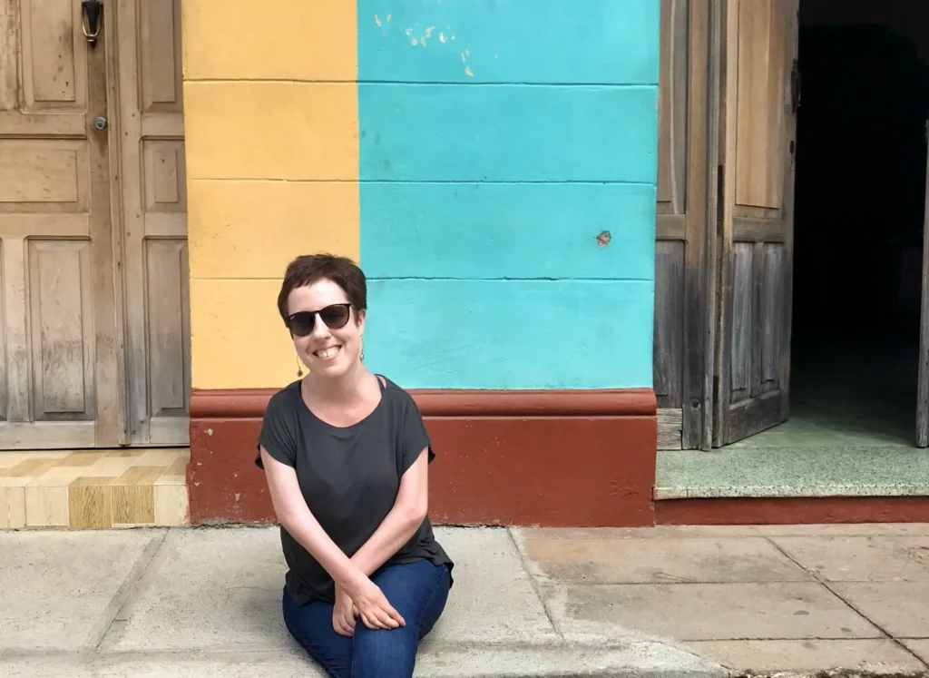 A woman wearing sunglasses sitting on a sidewalk against a colorful wall divided into yellow and turquoise sections, with wooden doors beside her. She is smiling, and the setting has a warm, casual vibe.