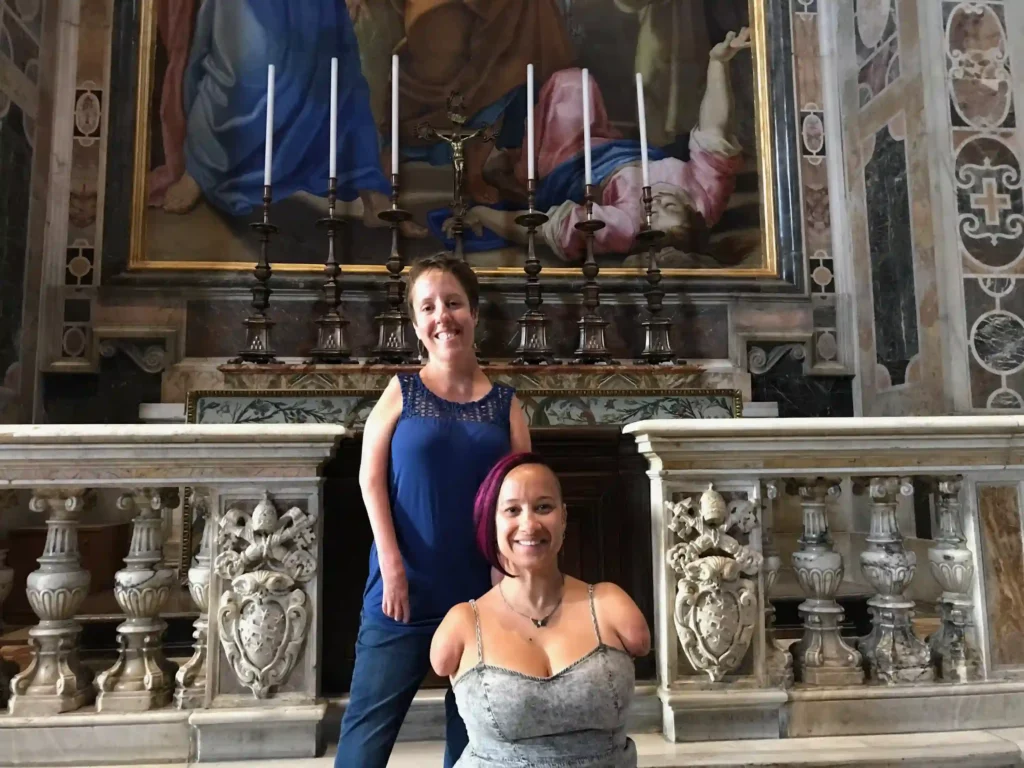 Two women pose together inside an ornate historic church. The woman standing in the back wears a blue sleeveless top and smiles warmly, while the woman sitting in the front, who has no arms, has short red hair, wears a gray sleeveless top, and smiles confidently. Behind them is a beautifully carved marble altar featuring intricate floral and emblematic details, along with decorative balustrades. The altar is adorned with tall metal candlesticks holding white candles, and above it, a large religious painting depicting biblical figures in vibrant colors dominates the background. The surrounding walls are intricately detailed with marble inlays and artistic motifs, adding to the grandeur of the setting.