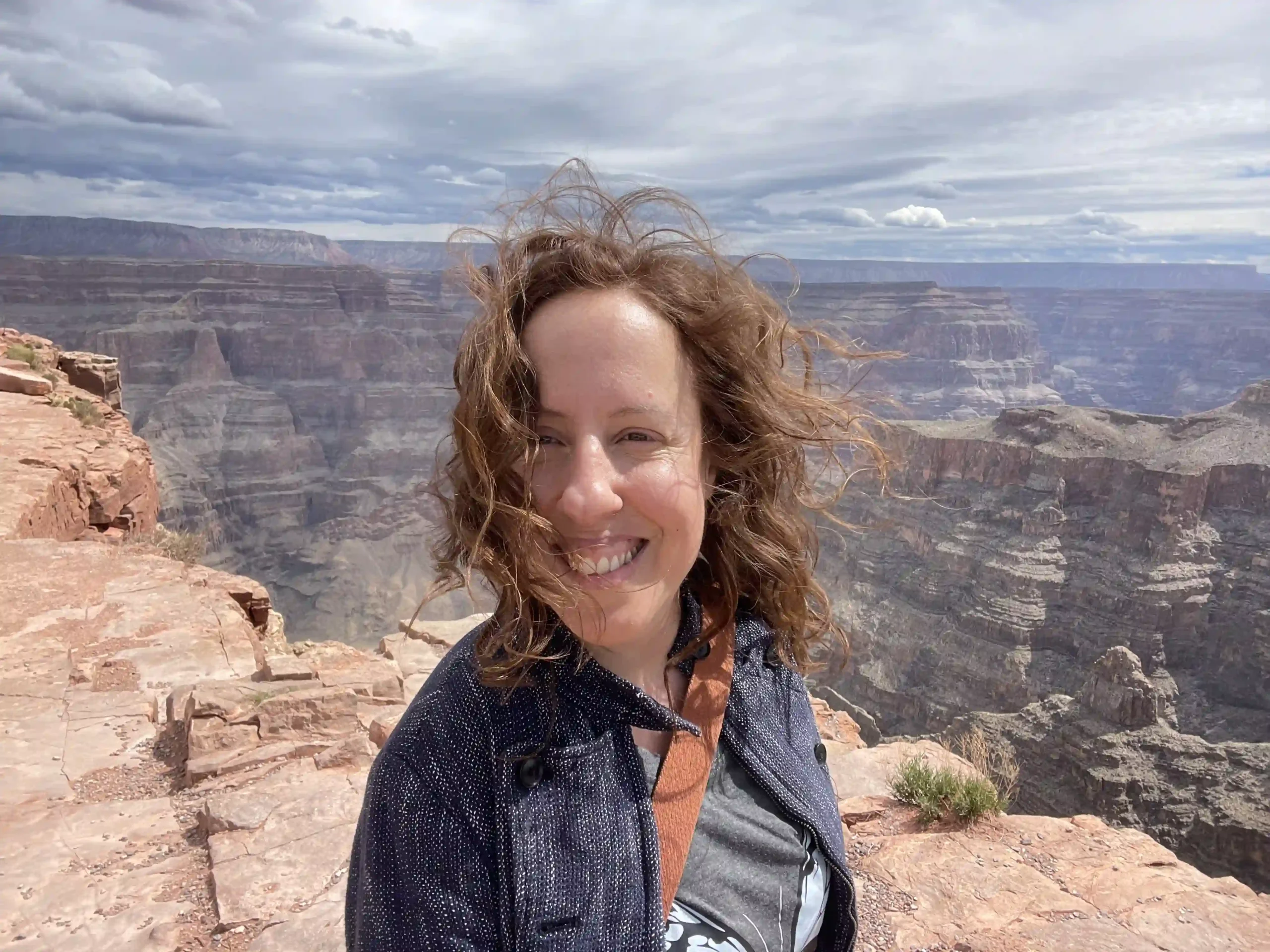 A smiling woman with curly hair standing on the edge of the Grand Canyon. Her hair is windswept, and the vast canyon landscape with rocky formations and cloudy skies fills the background.