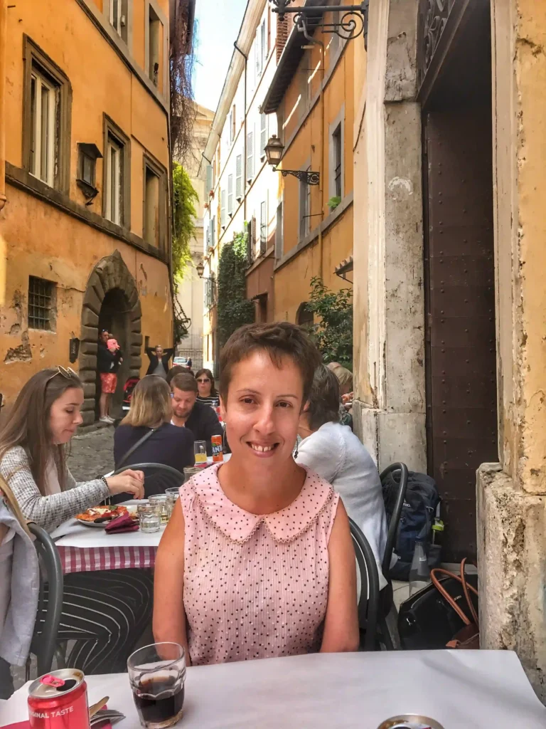 A woman sitting at an outdoor café in a picturesque alleyway with colorful buildings. She is smiling at the camera while dining, with other patrons in the background.