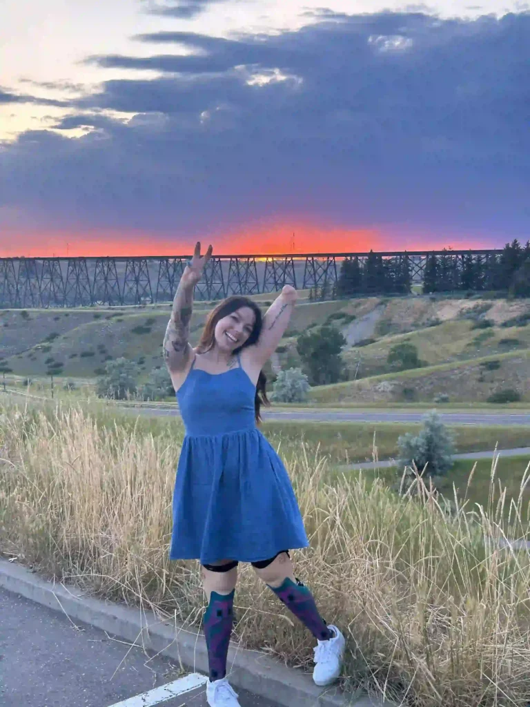 A young woman standing outdoors, smiling joyfully with arms raised in a peace sign gesture. She is wearing a sleeveless blue dress, colorful knee-high socks, and white sneakers. Behind her, a vibrant sunset paints the sky in shades of orange, pink, and purple, contrasting with dark clouds. A trestle bridge spans the background over rolling hills and grassy fields, adding depth to the scenic view. Tall grasses frame the foreground, enhancing the natural setting.