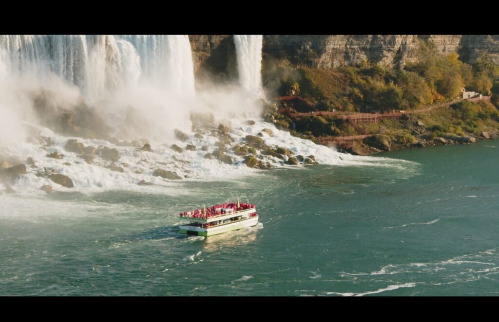 Tourist boat approaching the base of Niagara Falls with cascading waterfalls in the background, surrounded by rocky terrain and greenery.