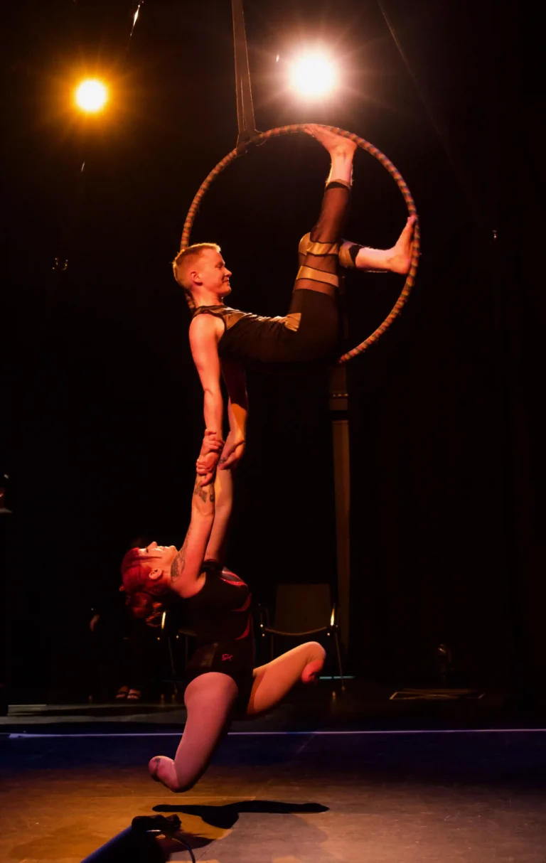 Onstage with low lighting, two white Disabled performers are in the air, on a suspended aerial hoop the size of an adult hula hoop at an average standing head height above the floor. Inside the hoop, Maxime sits side-profile, one leg up, leaning back into the curve of the hoop. Their arms hang straight down and holding onto them is Erin, a double below knee amputee. Erin is suspended, wearing no prosthetic legs, back arched like a crescent moon, and legs split apart. The two artists are smiling happily, celebrating their Queer Disabled Love. Photography: Jay Middaugh