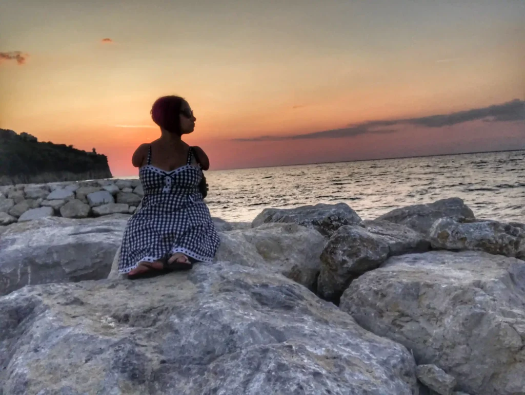 A woman with short hair and sunglasses is sitting on large rocks by the sea, wearing a black and white checkered dress. She is gazing towards the horizon where the vibrant orange and pink hues of the sunset blend with the blue of the sea. The background features a rugged coastline and a calm ocean.