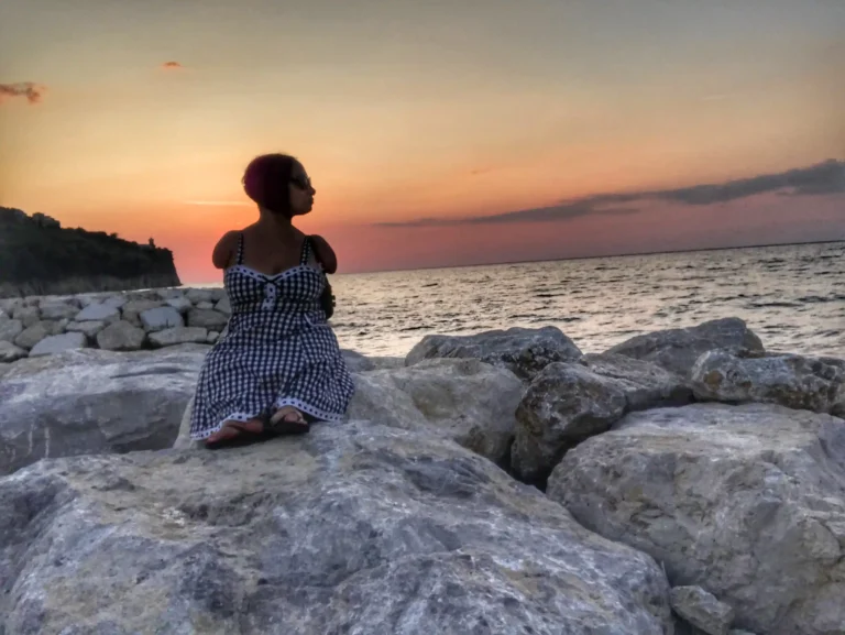 A woman with short hair and sunglasses is sitting on large rocks by the sea, wearing a black and white checkered dress. She is gazing towards the horizon where the vibrant orange and pink hues of the sunset blend with the blue of the sea. The background features a rugged coastline and a calm ocean.