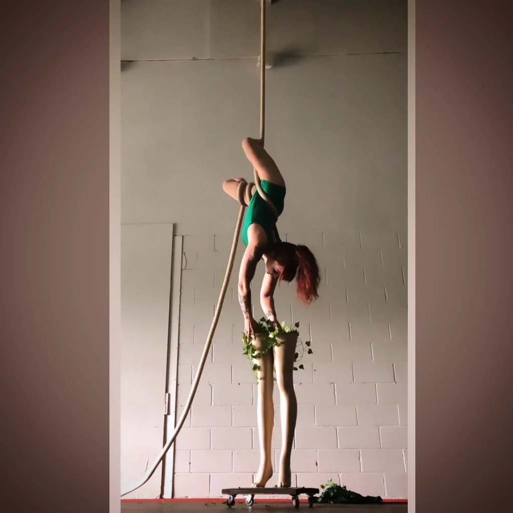 A covered rope is suspended from above. About half a story high, Erin, a white demifemme and double below knee amputee, is wrapped with the rope and upside down, reaching down to the mannequin legs below. The mannequin legs are open at the top and on a platform with wheels. Fake green plants and vines flow out of the top of the legs.