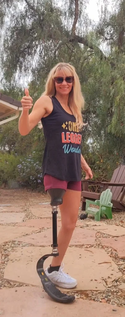 A woman standing outdoors with a prosthetic leg, smiling and giving a thumbs up, wearing a tank top that says "One-Legged Wonder."