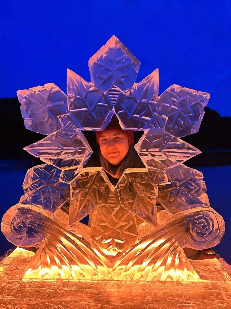 Person posing behind a detailed ice sculpture shaped like a large snowflake, with intricate patterns and a glowing orange light illuminating the base, set against a deep blue evening background.
