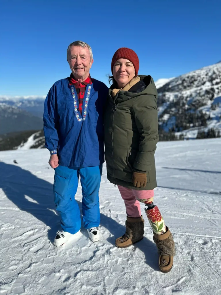 Two people standing on a snowy mountain with clear blue skies in the background. On the left, an older man wearing a blue jacket with decorative trim and blue pants stands smiling. On the right, a woman wearing a green jacket, red beanie, pink pants, and fur-lined boots smiles as well. The woman's prosthetic leg features a colorful floral design
