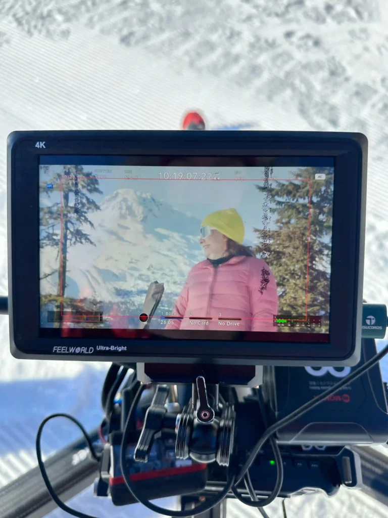 A close-up photo of a camera monitor (FEELWORLD Ultra-Bright 4K) displaying a scene being filmed outdoors in a snowy environment. The screen shows a person wearing a pink jacket, yellow beanie, and reflective sunglasses with snow-covered trees and mountains in the background. The camera rig and various cables are visible below the monitor, with a clear snowy surface in the backdrop.