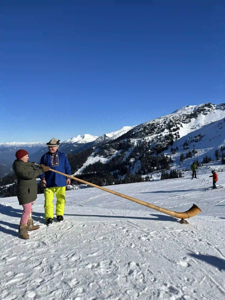 Two people standing on a snowy mountain slope, one playing a long wooden Alphorn, a traditional Swiss instrument. The scene is set against a backdrop of stunning snow-covered peaks and a clear blue sky, with skiers visible in the distance.