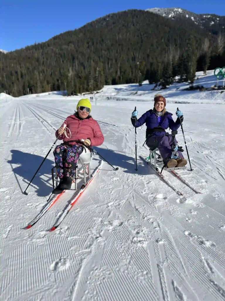 Two individuals sitting in adaptive ski chairs on a snowy trail with ski poles in hand, surrounded by a scenic mountain landscape under a clear blue sky