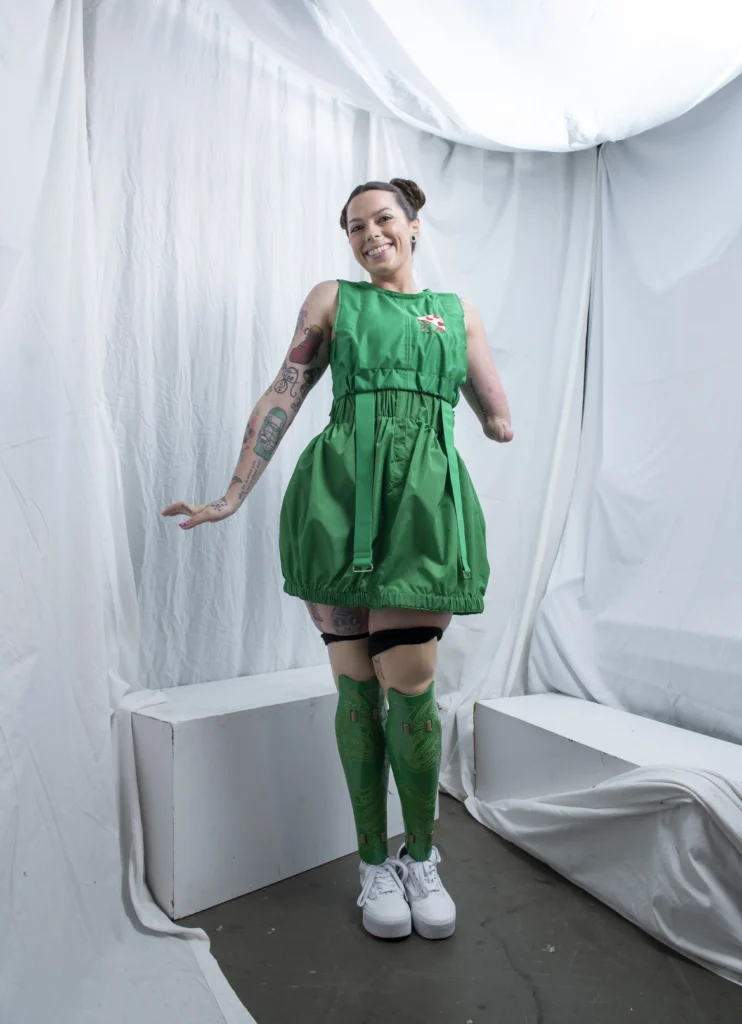 A person posing gracefully in a vivid green dress with matching green prosthetic legs. They are standing on a white set with draped fabric and geometric blocks, exuding a joyful and confident demeanor. Y Hanson Photography Fashion Nomads, Vancouver Fashion Week, 2024