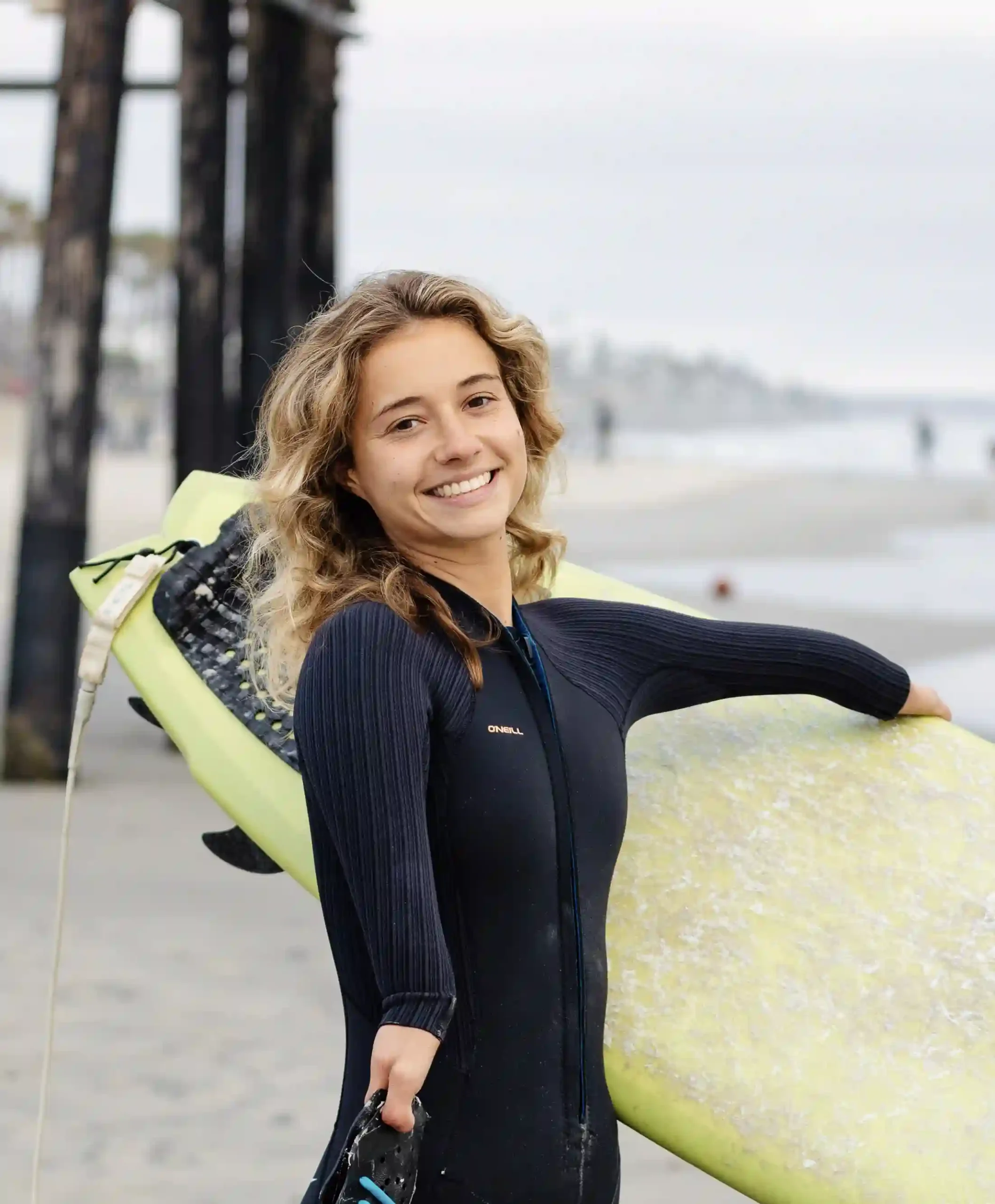 Young woman with curly blonde hair smiling and holding a yellow surfboard under her arm. She is wearing a black wetsuit and standing on a beach with the ocean and a pier visible in the background.