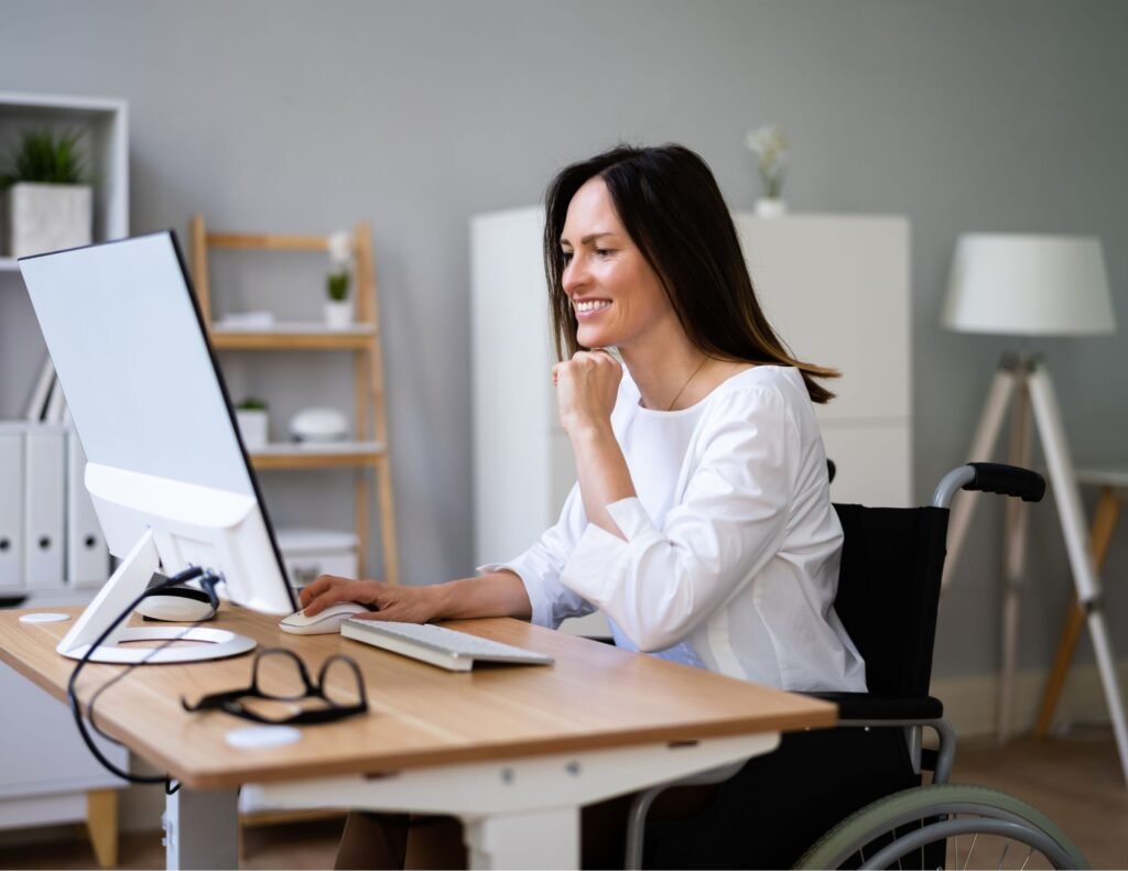 A woman in a wheelchair is sitting at a desk, smiling while working on a computer in a bright and modern home office setting. The desk has a pair of glasses and other office supplies on it, and the background includes shelves and a lamp, adding a cozy and professional ambiance.