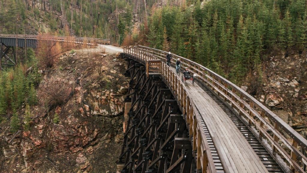 A scenic view of a wooden trestle bridge crossing a rocky canyon surrounded by evergreen forest. The bridge has sturdy railings, and there are cyclists riding across, enjoying the outdoor adventure. Accessible Biking at Myra Canyon Trestles, Kelowna