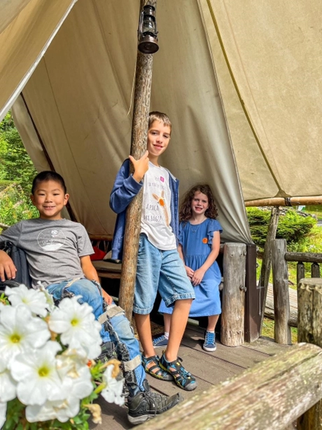 Three children standing and sitting near a wooden tent structure in an outdoor setting, surrounded by green foliage. The child on the left is seated and wearing a gray shirt, the child in the middle is standing in a blue jacket and denim shorts, and the child on the right is seated in a blue dress. A cluster of white flowers is visible in the foreground