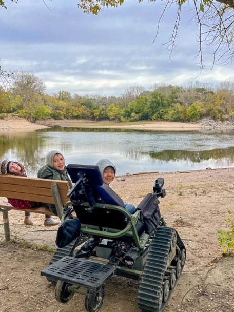A group of three children sitting by a scenic lake, surrounded by trees with autumn foliage. Two people are on a bench smiling towards the camera, while the third is in a rugged all-terrain wheelchair designed for outdoor use, also smiling. The overcast sky adds a calm, serene atmosphere to the scene.