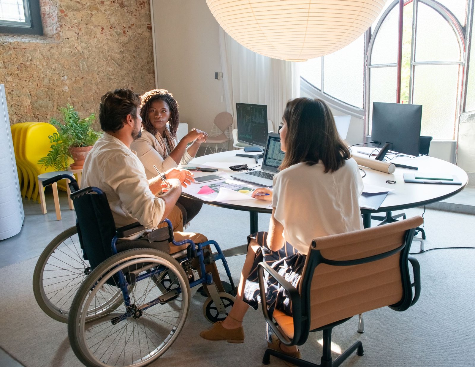 A diverse group of professionals engaged in a discussion around a round table in a modern office space. One participant is a man in a wheelchair, contributing ideas as others listen attentively. The workspace features large arched windows, natural light, and an inviting atmosphere with plants and minimalistic decor, emphasizing collaboration and inclusivity.