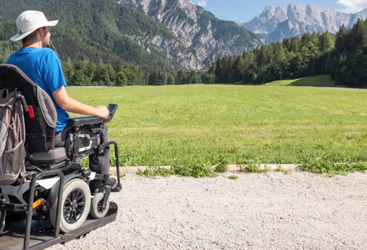 A man in a power wheelchair wearing a white sun hat and blue shirt enjoys a scenic view of a lush green meadow surrounded by tall pine trees, with majestic mountains in the background under a clear blue sky.