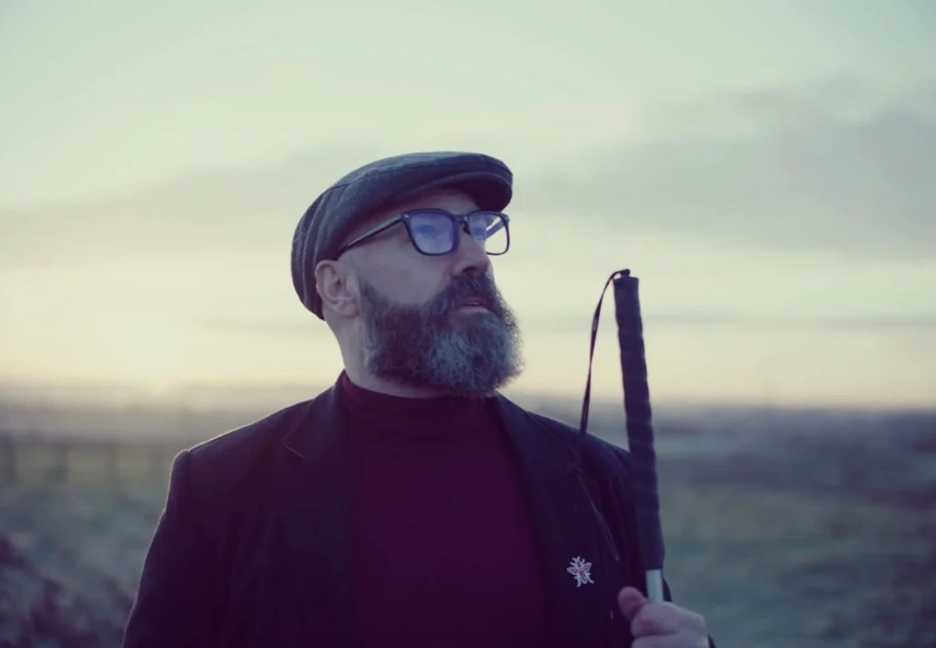 Man with a beard and glasses wearing a flat cap and dark clothing, standing outdoors at sunset, holding a white cane