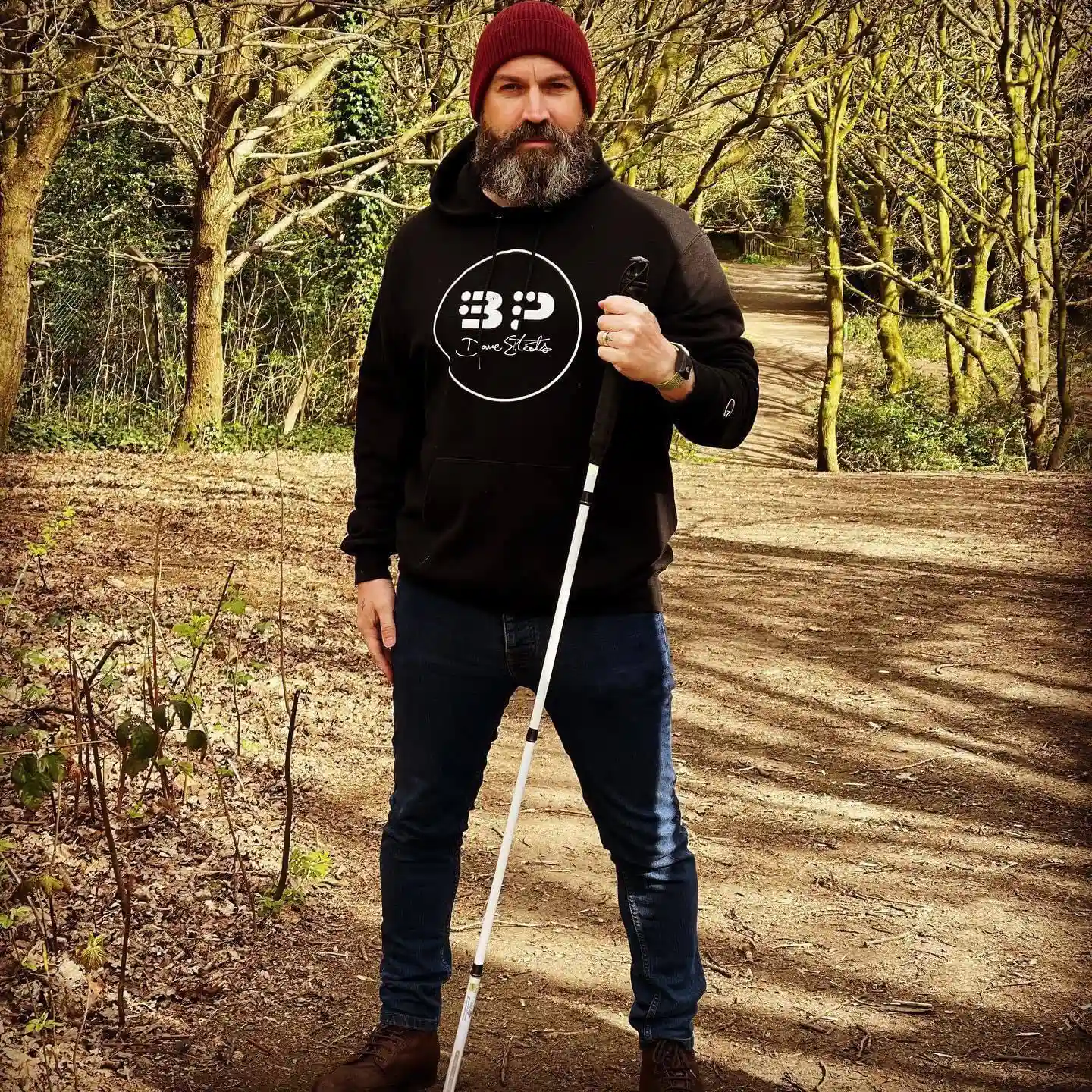 Man with a beard wearing a beanie, dark hoodie, and jeans, holding a white cane while standing on a wooded trail surrounded by trees