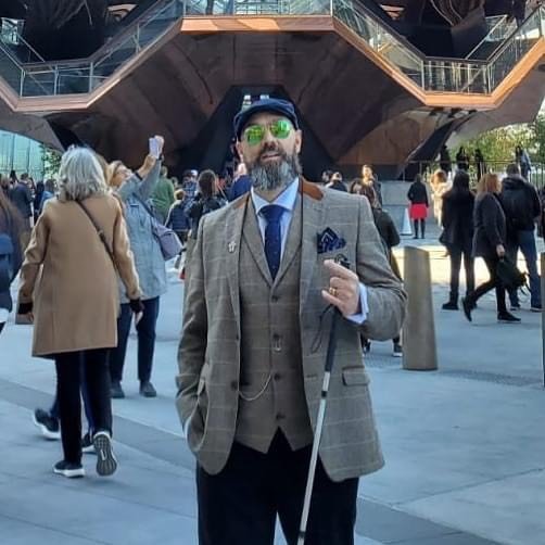 Man wearing a stylish brown checkered suit, navy tie, and pocket square, holding a cane and standing in front of a modern architectural structure with a crowd in the background. He has a gray beard, sunglasses, and a cap, exuding confidence.