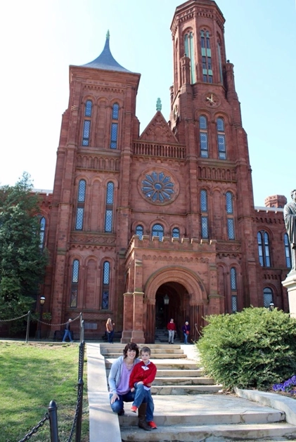 Two people sitting on the steps leading to the Smithsonian Institution Building, also known as the Castle, a historic red sandstone structure with gothic architecture, located in Washington, D.C. The background features tall towers, arched windows, and a large circular stained-glass window.