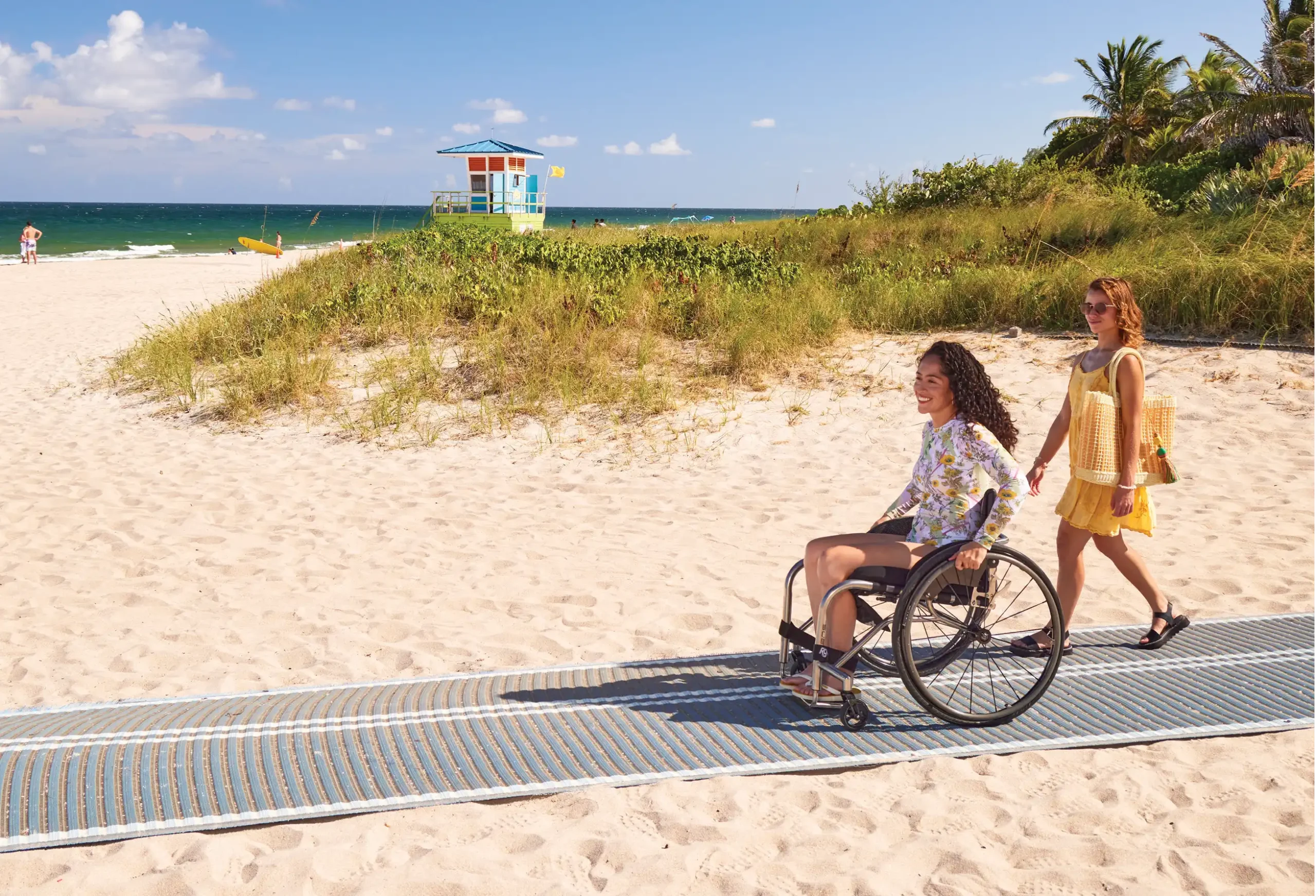 Two women enjoying a sunny day at the beach, one is using a wheelchair and rolling along an accessible beach pathway, while the other walks alongside her in a yellow dress. In the background, there is a lifeguard tower, greenery, and the ocean under a blue sky
