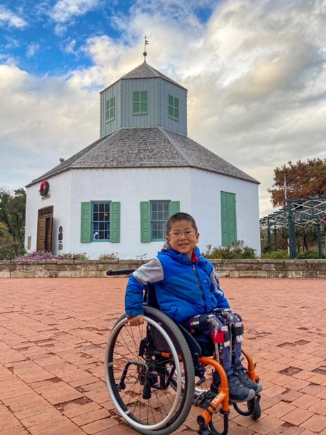 A young boy in a blue jacket, sitting in a wheelchair, smiles in front of a historical white octagonal building with green shutters and a small tower on top. The scene is set outdoors on a paved area under a partly cloudy sky, with greenery and holiday decorations visible in the background.