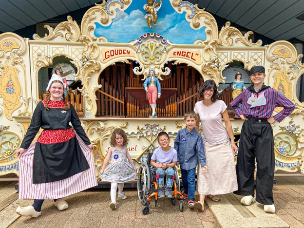 A group photo in front of a decorative organ labeled "Gouden Engel." The group consists of six individuals, including two children, one using a wheelchair, standing alongside adults in traditional Dutch clothing and casual attire. The scene is cheerful, with vibrant colors and an ornate backdrop showcasing Dutch cultural elements.