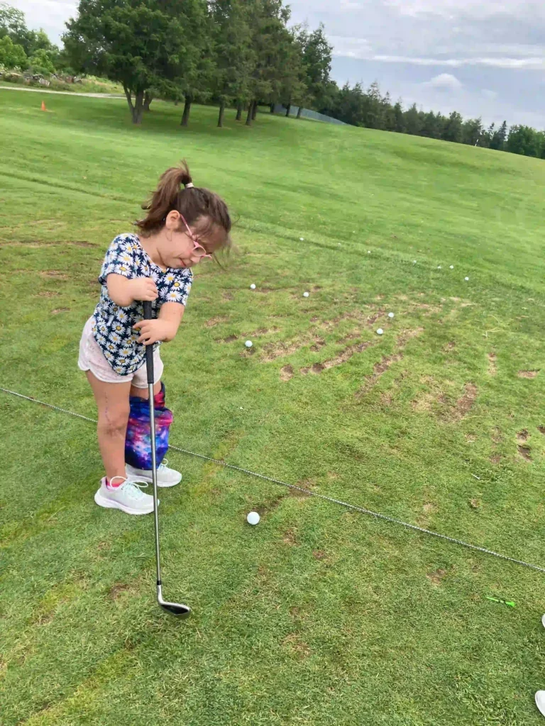 A young child with glasses stands on a green golf course, holding a golf club and preparing to hit a golf ball. She is wearing a blue floral shirt, pink shorts, and white sneakers. A colorful bag hangs by her side. The background features trees and a bright, open field with scattered golf balls on the ground.