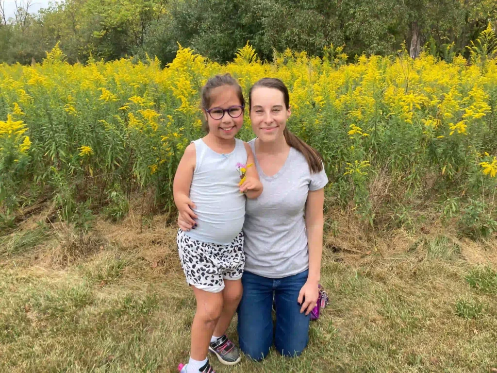 A woman and a young girl are posing outdoors in front of a vibrant field of yellow flowers. The woman is kneeling on the grass, wearing a light gray T-shirt and jeans, with her hair tied back in a ponytail. The girl is standing next to her, smiling, and wearing glasses, a white tank top, and leopard print shorts. Both appear happy and are embracing warmly, with greenery and trees in the background.
