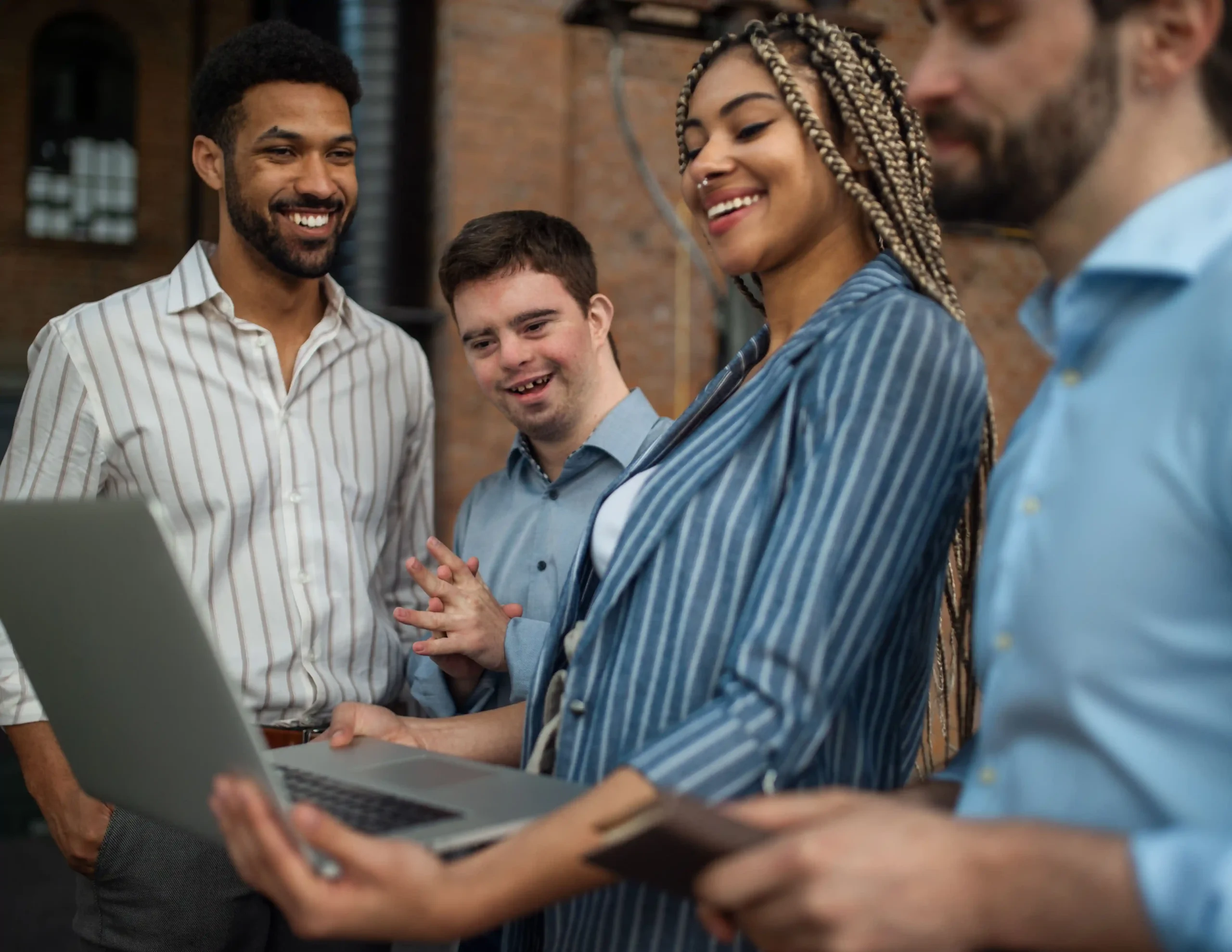 A diverse group of professionals smiling and collaborating around a laptop. One of the individuals in the group has Down syndrome, showcasing inclusivity in the workplace. They are standing in an open office setting, engaging in discussion