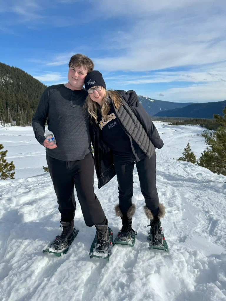 A man and a woman smiling while snowshoeing on a snowy mountainside under a bright blue sky. The man is wearing a gray long-sleeve shirt and holding a water bottle, while the woman is dressed warmly in a black jacket, black pants, and a beanie with fur-lined boots. They appear happy, standing close together with scenic snowy terrain and forested mountains in the background.