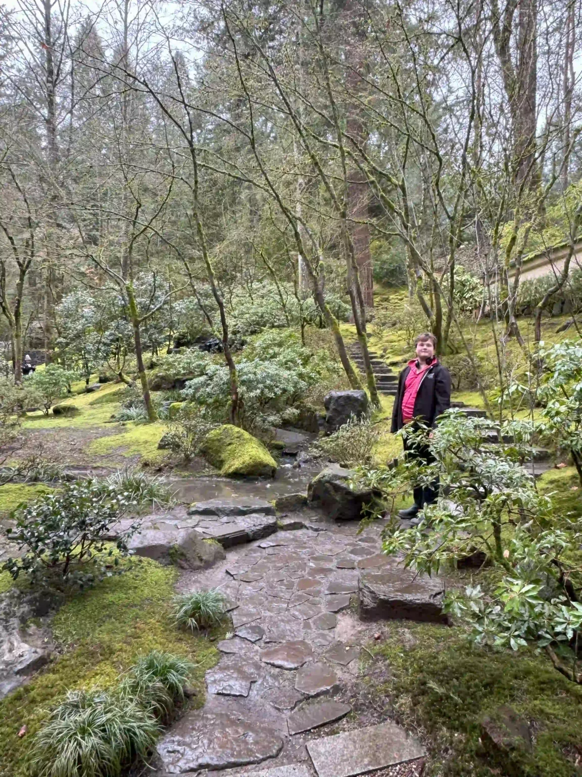 A serene garden scene at the Portland Japanese Garden, featuring lush green moss covering the ground, stone pathways, and a variety of trees and shrubs. A person wearing a red shirt and black jacket stands near the pathway, surrounded by the tranquil natural setting.