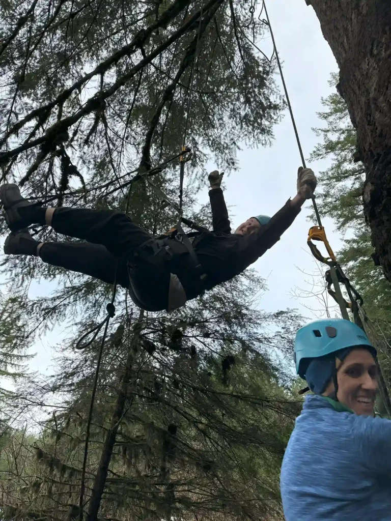 Two people engaging in recreational tree climbing in a forested area. One person, wearing black climbing gear, is suspended in the air by ropes, smiling and reaching out. The other person, wearing a blue helmet and jacket, is on the ground holding a climbing rope and smiling. The background features tall trees and a cloudy sky