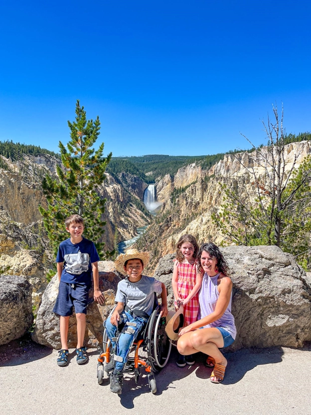 A family of four posing in front of a scenic canyon with a waterfall in the background on a sunny day. The group includes a boy, a girl, a woman, and a person in a wheelchair wearing a straw hat. The backdrop features rugged cliffs, a vibrant blue sky, and lush greenery.