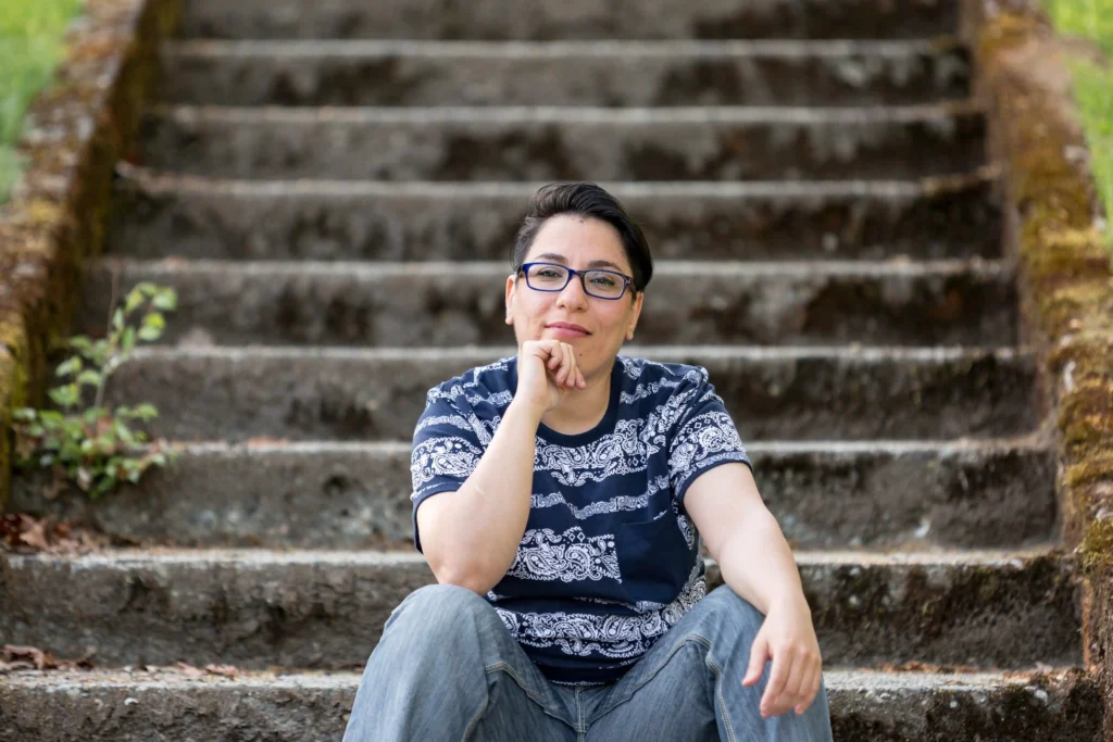 A person with short dark hair, wearing glasses, a patterned blue t-shirt, and jeans, sitting casually on outdoor stone steps. They are resting their chin on their hand and smiling slightly. The background features moss-covered edges and greenery.