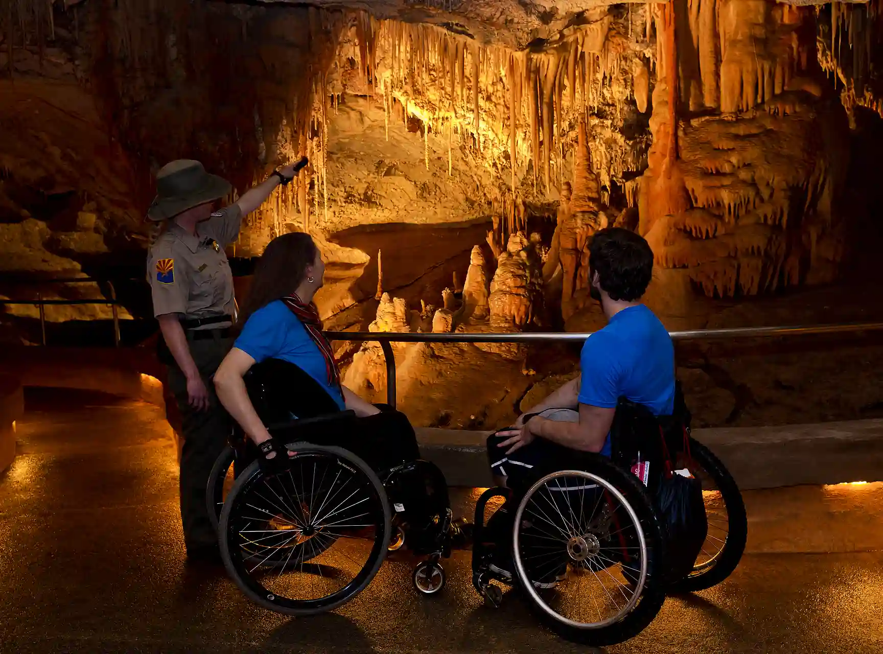 Three people exploring an illuminated cave with stalactites and stalagmites, including a park ranger and two visitors in wheelchairs, observing the geological formations