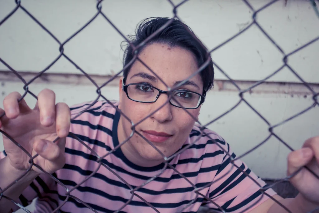 Person wearing glasses and a pink striped shirt, looking through a chain-link fence with a thoughtful expression, their hands lightly gripping the fence.