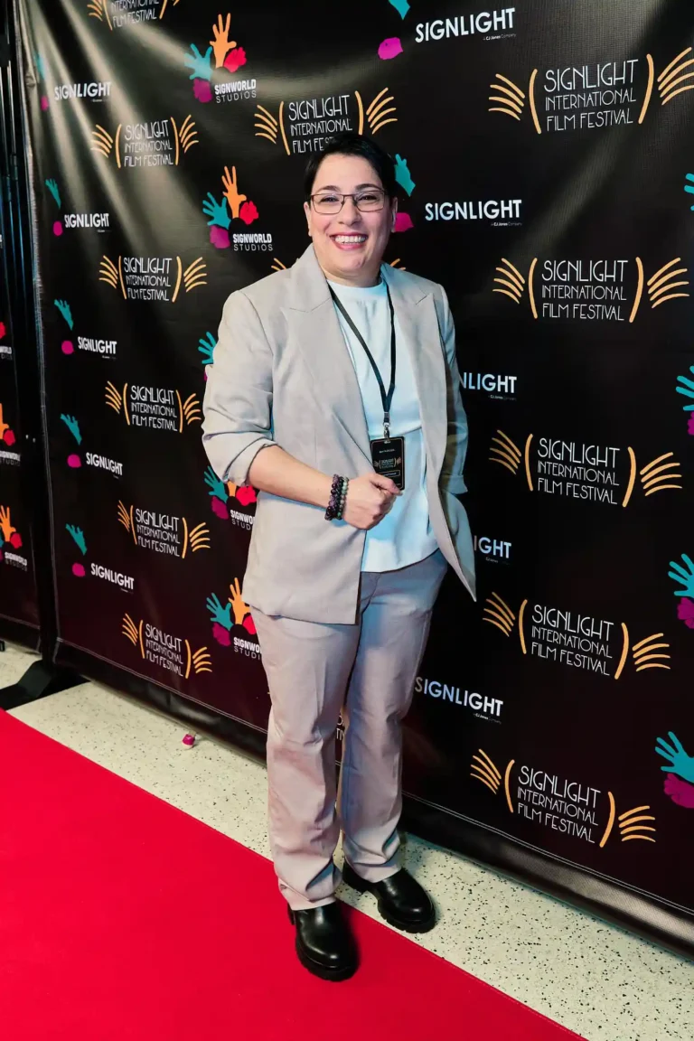 A person with short dark hair and glasses stands on a red carpet at the SignLight International Film Festival. They are dressed in a light beige suit, a white top, and black shoes, smiling broadly. The backdrop behind them has the festival's colorful logo and branding.